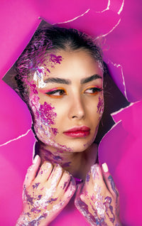 A woman with art makeup peeks through pink paper.